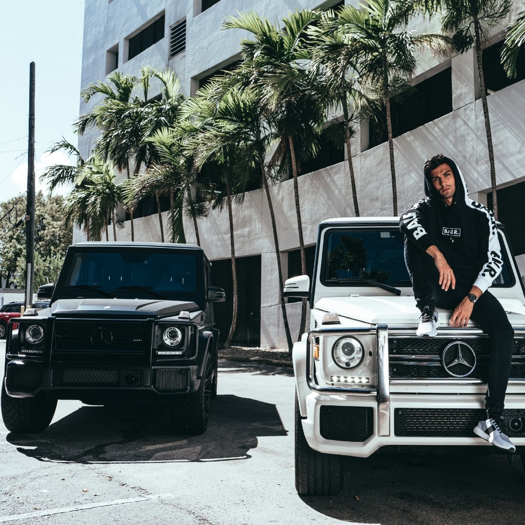 A young man in casual streetwear sitting on the hood of a white Mercedes-Benz G-Class SUV, with a black Mercedes-Benz G-Class SUV parked beside him, in front of a modern building with tall windows and palm trees.
