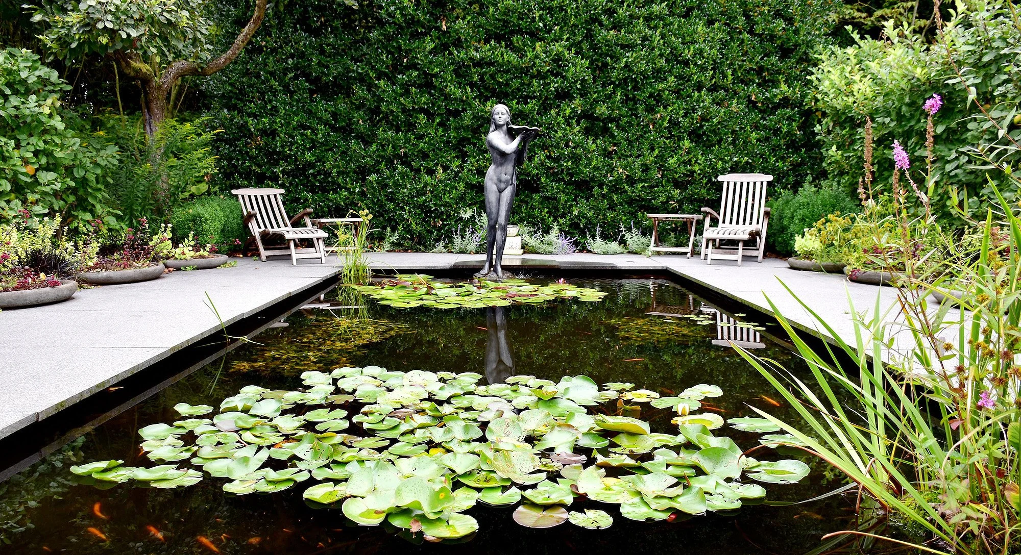 A rectangular pond surrounded by a concrete path with water lilies and small fish. In the background, a statue of a woman playing a flute, flanked by benches and lush greenery.