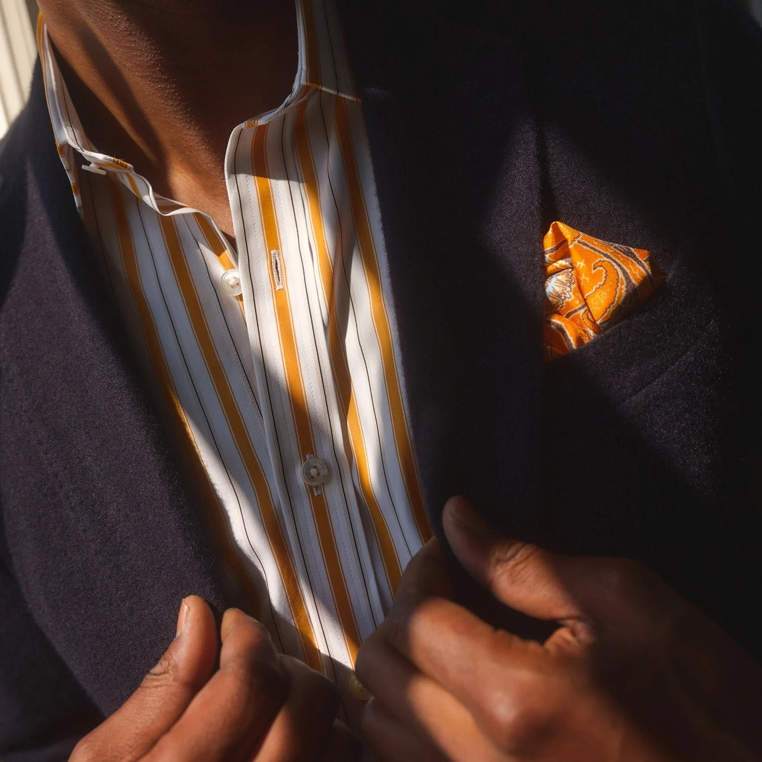 Close-up of a man in a dark blazer pulling it back to reveal a striped shirt with orange, white, and gold colors and a matching pocket square, with a focus on the fabric details.