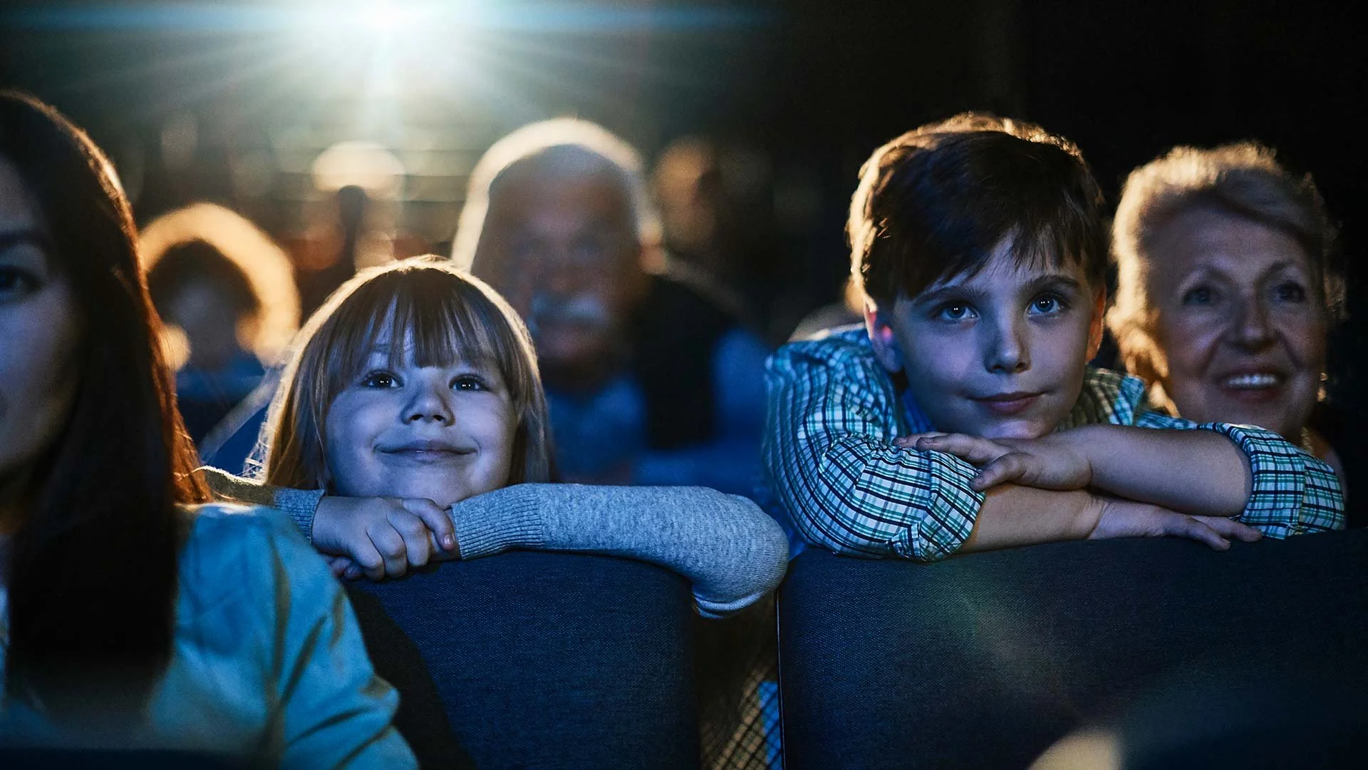 Children and adults seated in a dark theater, illuminated by the glow of a screen, watching a movie or performance with expressions of joy and interest.