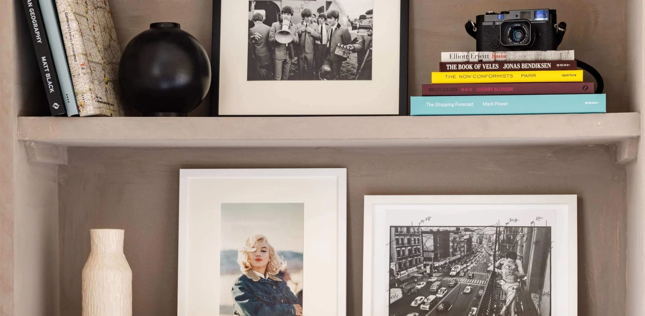 Decorative shelf displaying a black round vase, a black-and-white historical photograph, a camera, a stack of books titled 'Kids', 'The Book of Veles', 'The Non-Conformists', 'Cherry Blossom', 'The Shipping Forecast', and a framed photograph of a woman with blonde hair, and a framed black-and-white cityscape photo.