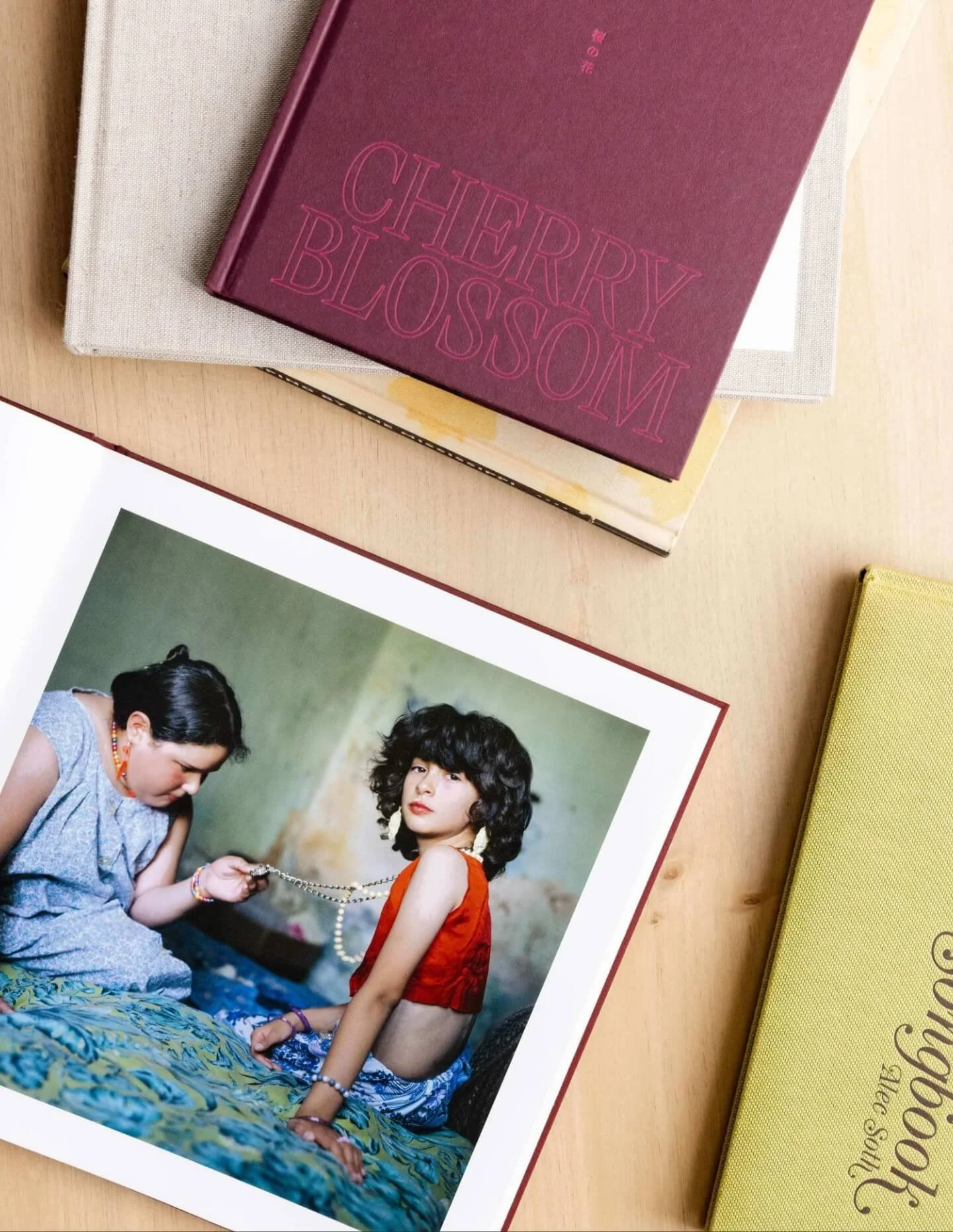 A printed photograph of two women, one with dark, curly hair and a red top, sitting on a bed, holding a necklace, and another with dark, straight hair, looking down, wearing a sleeveless dress. The photo is placed on a light wooden surface along with several closed books or journals, including one titled "Cherry Blossom" in pink, and another in a yellow cover.