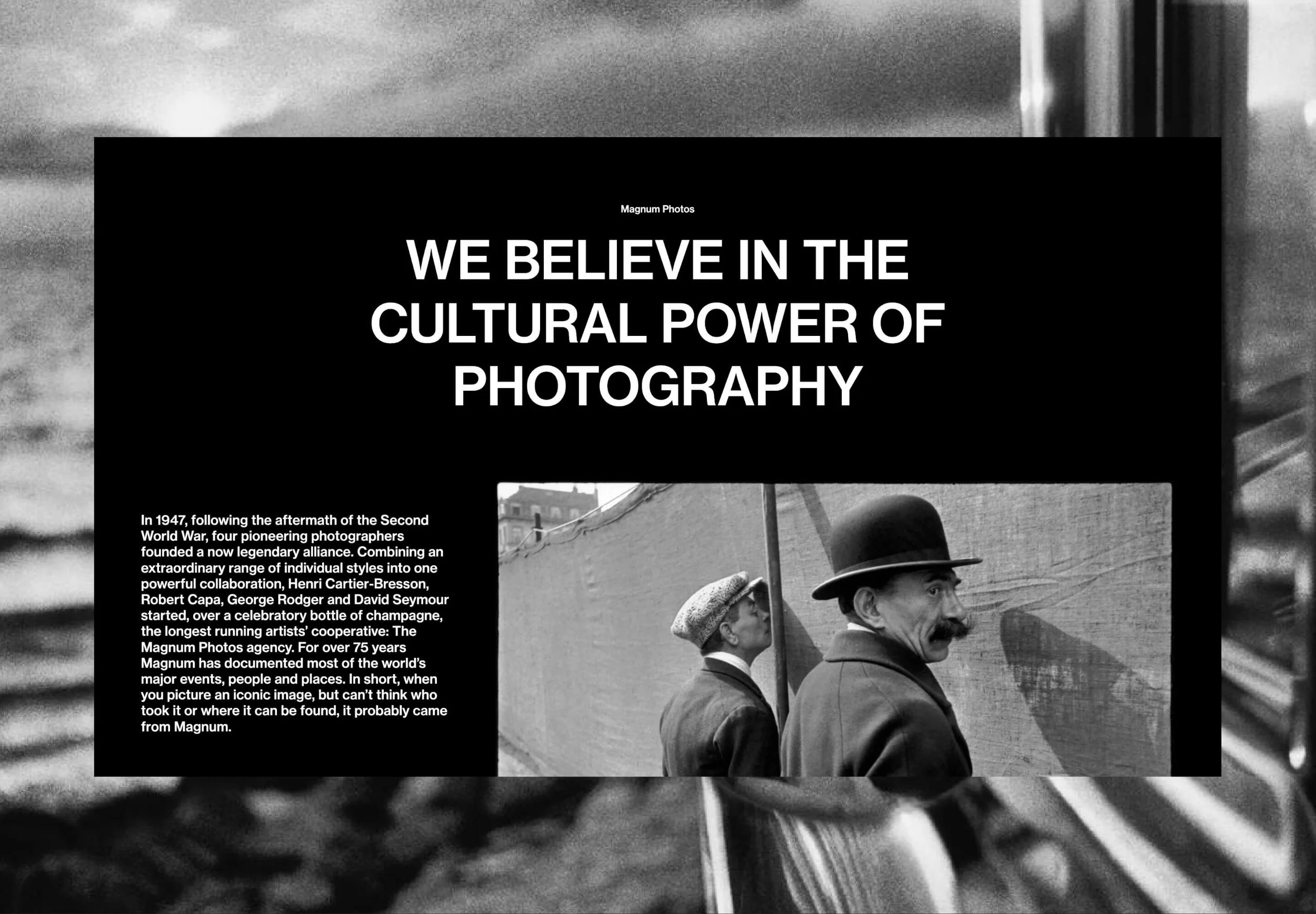 Black and white photo of two men, one wearing a bowler hat and the other a flat cap, standing beside a large wall with a cityscape in the background.