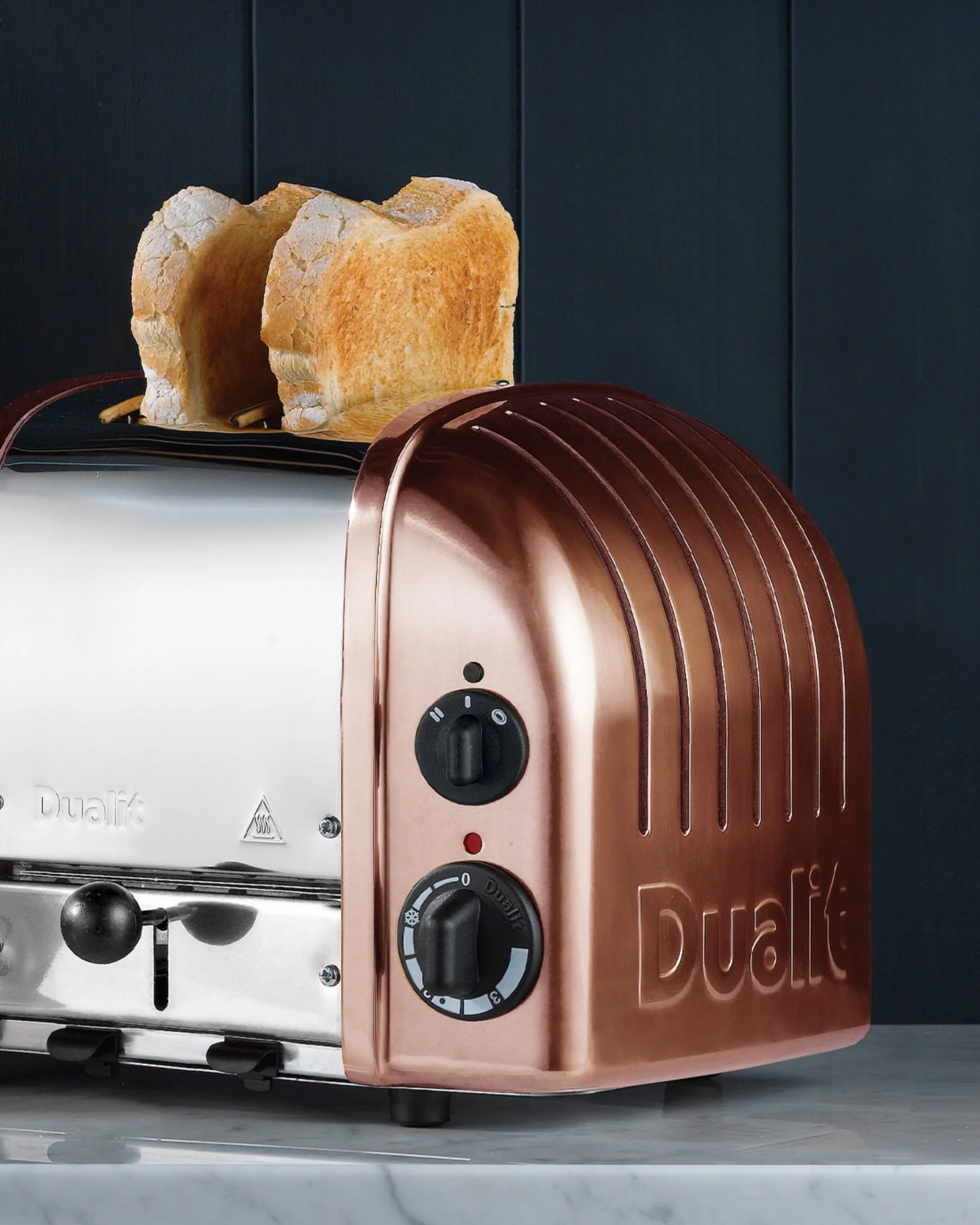 A toaster with two slices of toasted bread on top, placed on a marble countertop against a dark wall.