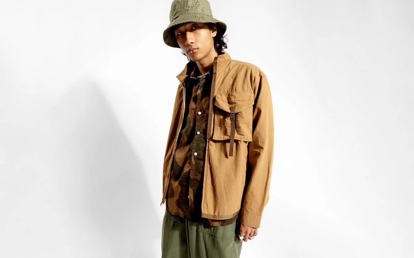 Young man wearing a camouflage bucket hat, brown jacket, and olive-green pants, standing against a plain white background.
