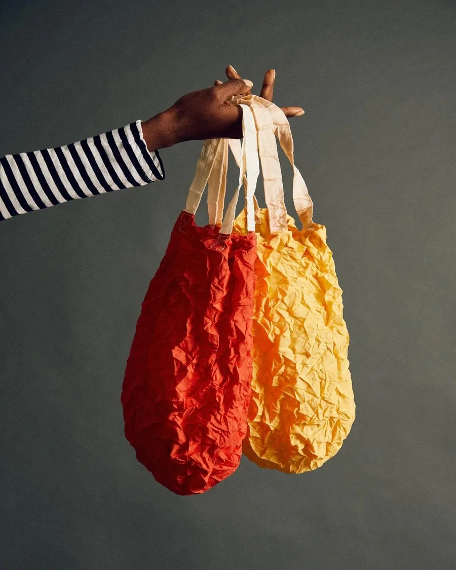 A person holding two crumpled reusable shopping bags, one red and one yellow, against a plain, dark background.