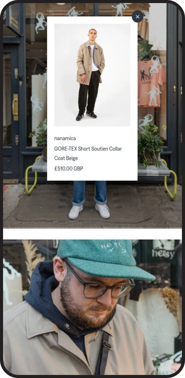 Urban outdoor scene with two people; one is obscured by a mobile advertising display featuring a model wearing a beige coat, black pants, and white sneakers, with details about the coat, priced at £510.00 GBP; the other person, visible in the lower part of the image, is a man wearing glasses, a green cap, a beige coat, and a dark hoodie.