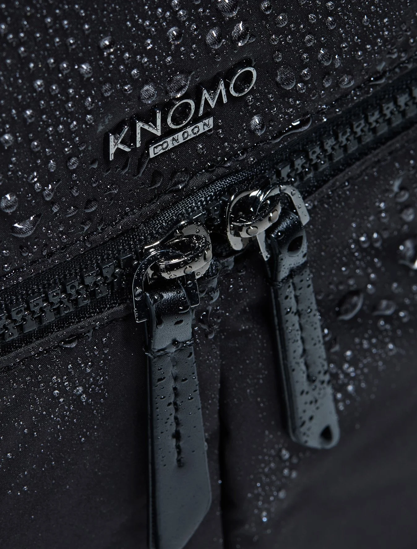 Close-up of a black Knomo backpack with water droplets and zippers.