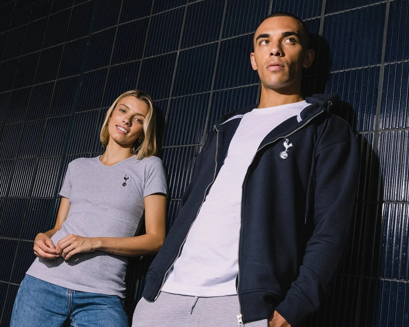 Two young adults, a woman and a man, leaning against a dark tiled wall, wearing Tottenham Hotspur sportswear.