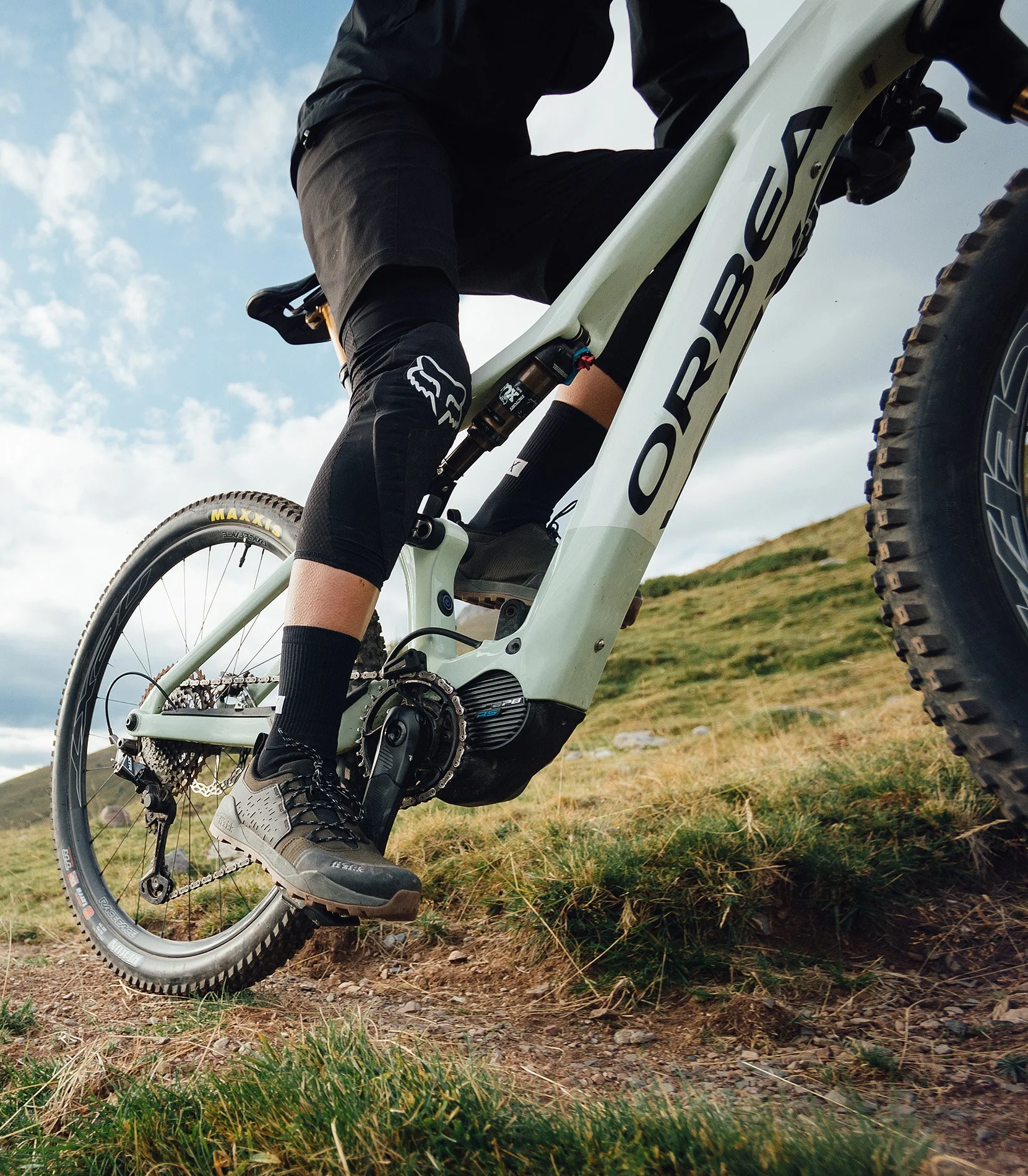 Close-up of a person riding an electric mountain bike on a grassy trail, wearing black gear and sneakers, with a cloudy sky in the background.