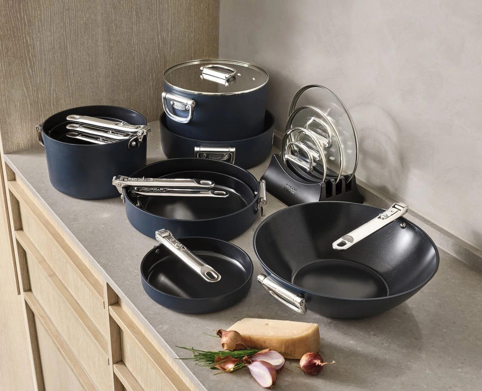 Set of black non-stick pots and pans with silver handles on a kitchen countertop, alongside a block of cheese, garlic, and herbs.