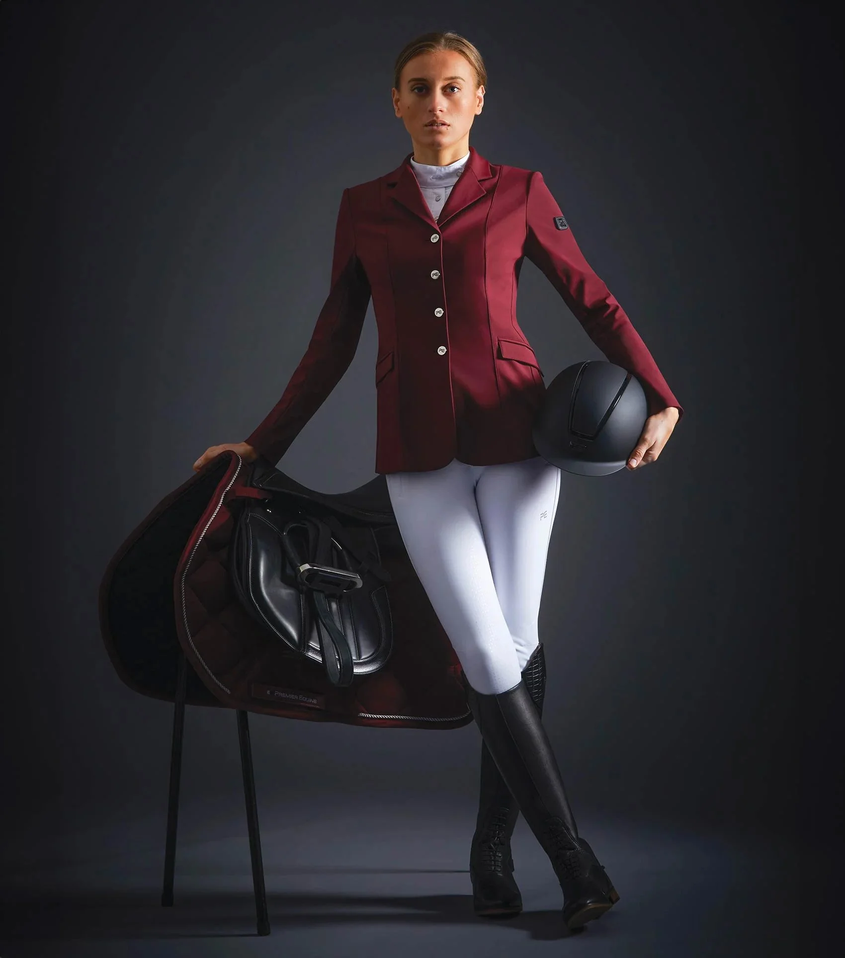 A woman wearing a maroon blazer, white riding pants, and black riding boots, holding a black riding helmet in one hand and standing next to a chair with a helmet and riding gear on it, against a dark background.