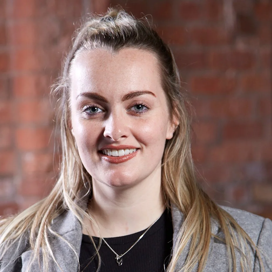 A young woman with long blonde hair, blue eyes, and fair skin, smiling in front of a brick wall.