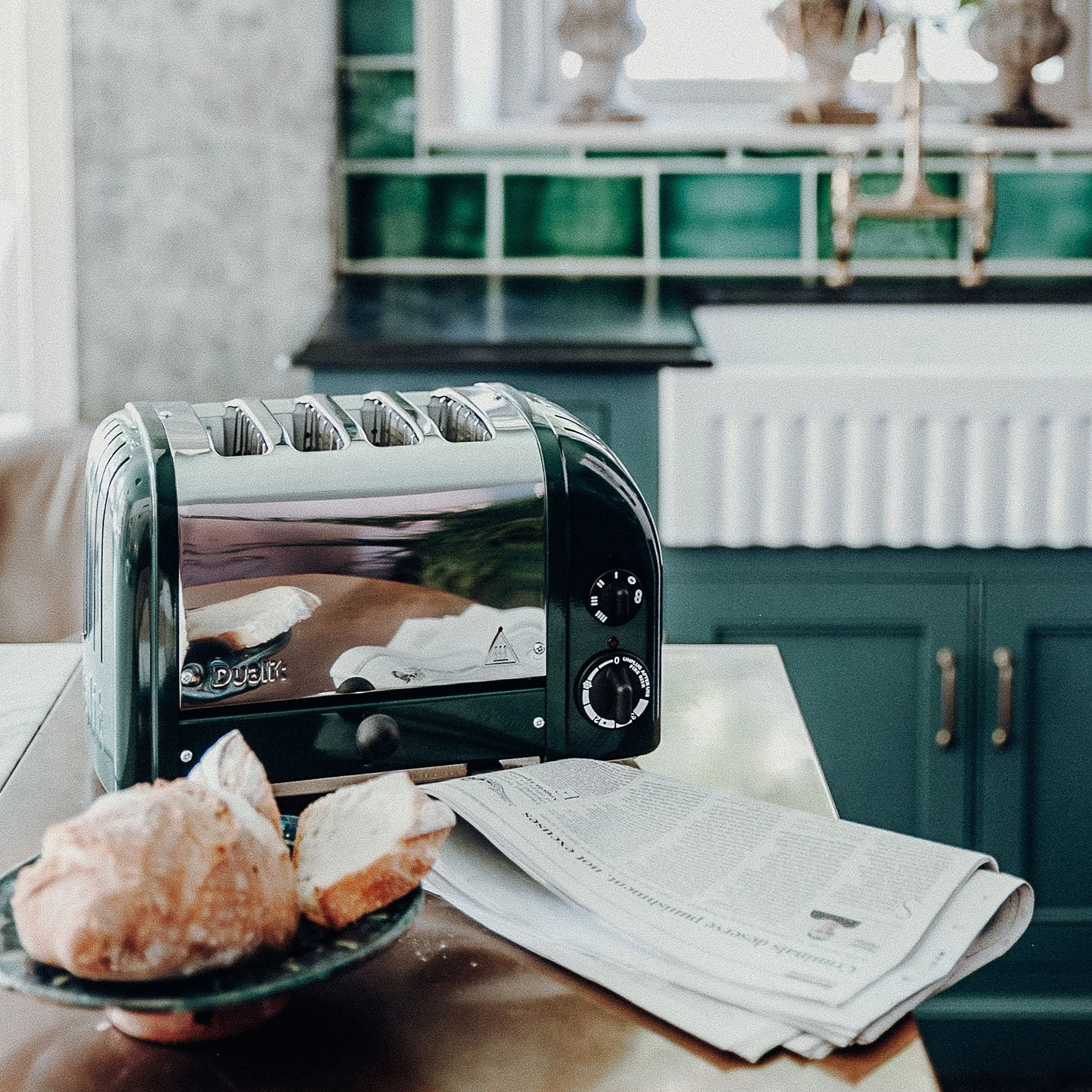 A modern black toaster with four slots on a kitchen counter, next to a plate of bread rolls and a folded newspaper, with green cabinets and a window in the background.