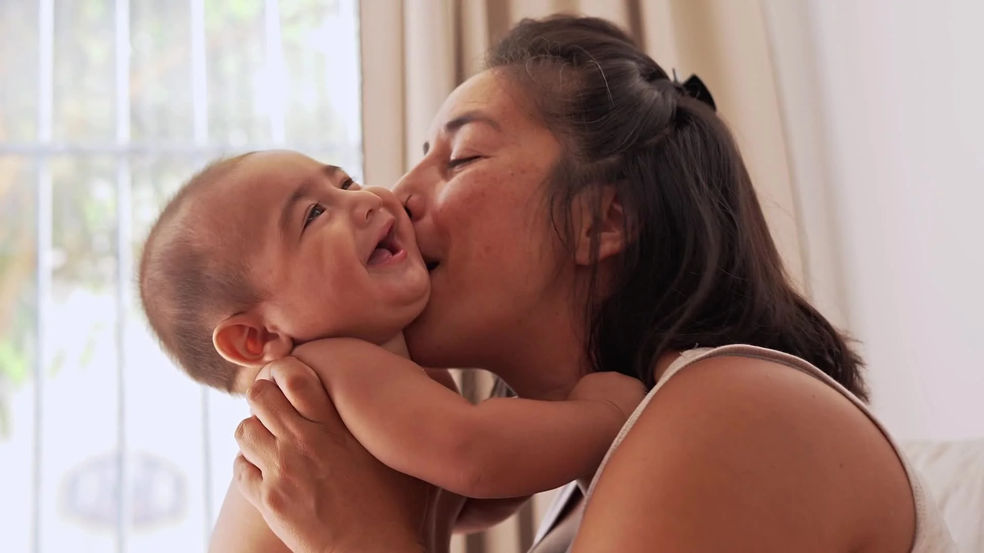 Mom hugs and kisses her smiling young son indoors with sunlight coming through the window.