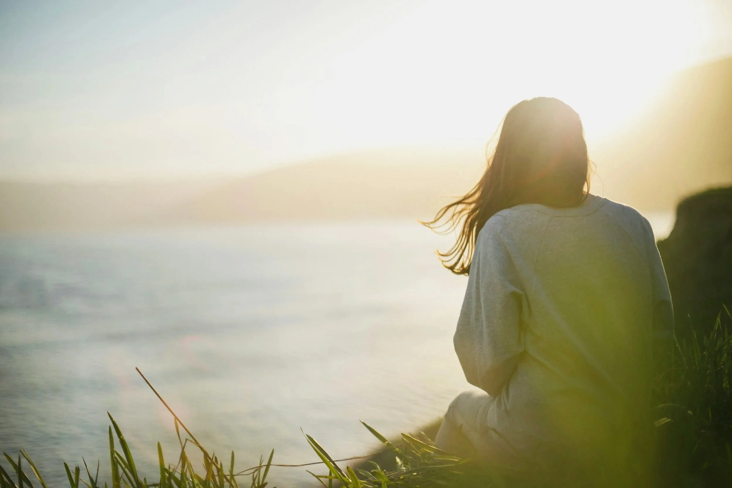 A person with long hair fishing by a lake or river during sunset, standing near tall grass.