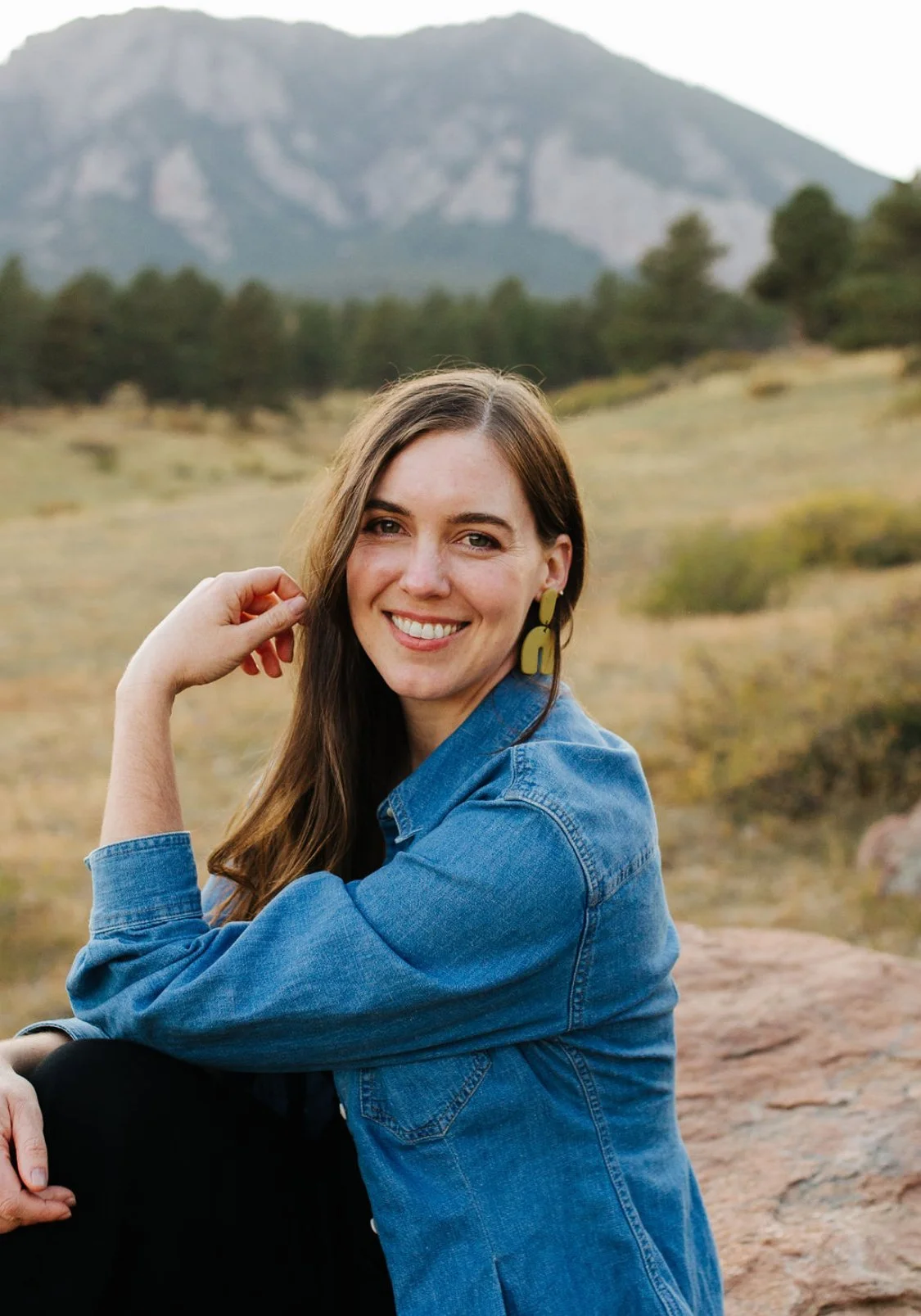 A woman with long brown hair smiling and sitting outdoors in a natural landscape with mountains and trees in the background.