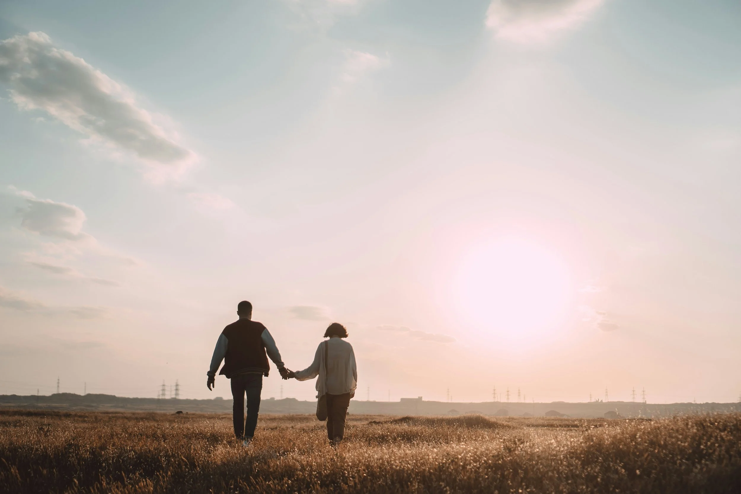 A couple walking hand in hand in a field at sunset, with power lines and a few clouds in the sky.