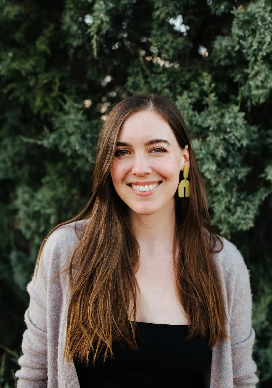 A woman with long brown hair and yellow earrings, smiling outdoors with greenery in the background.