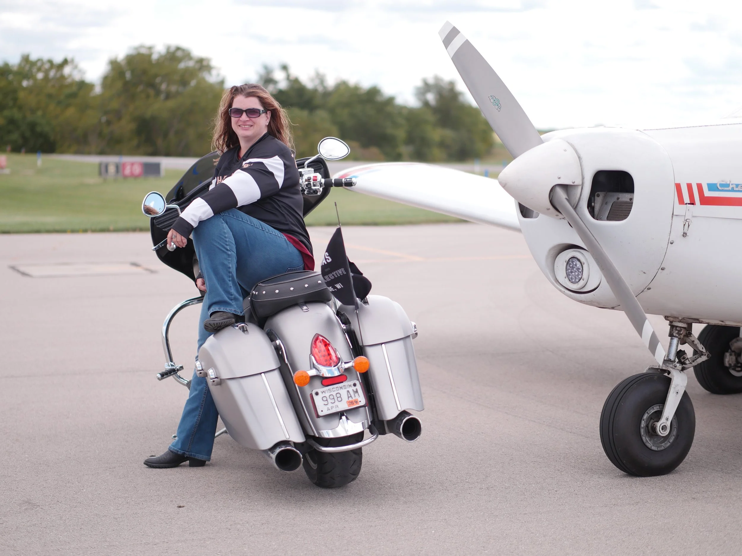 A woman sitting sideways on a motorcycle on an airport tarmac, smiling, wearing sunglasses, black and white jacket, blue jeans, with a small plane in the background.