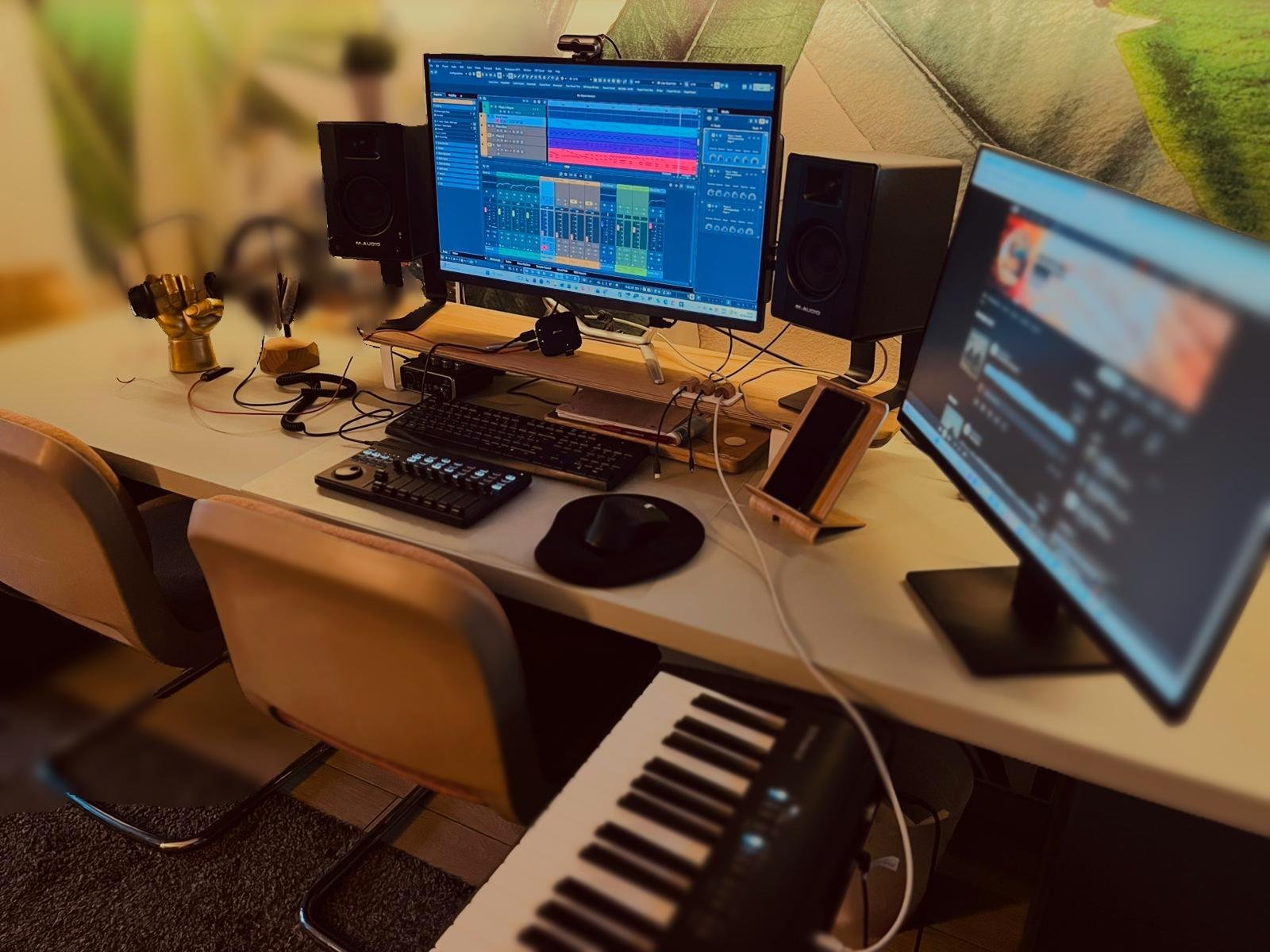 A music production workspace with two monitors displaying digital audio workstation software, connected to speakers, a MIDI keyboard, a control surface, a mouse, a keyboard, and a smartphone on a desk. There is also a small keyboard instrument in the foreground.