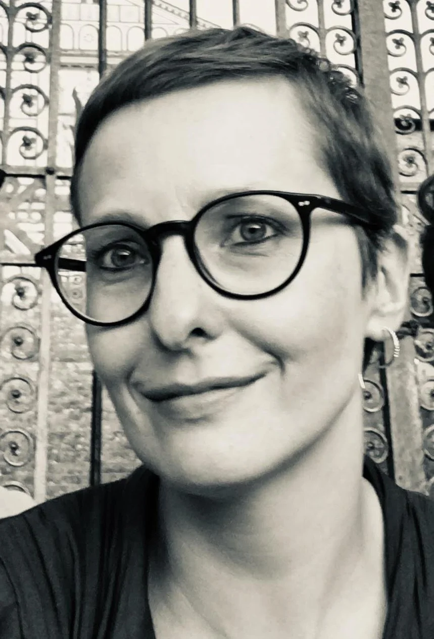 A woman with short hair, wearing glasses and earrings, smiling at the camera with a decorative iron fence in the background.