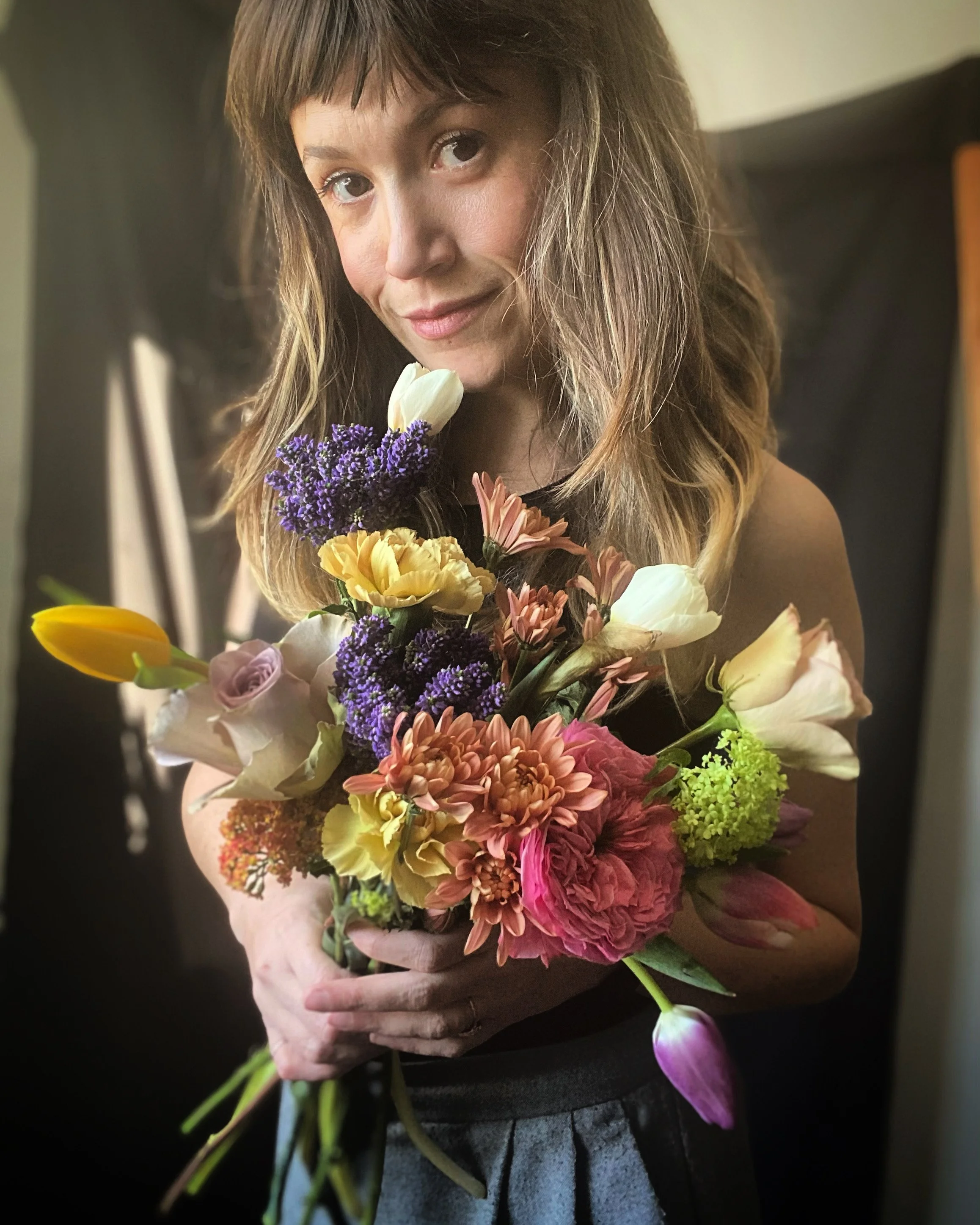 A woman holds a colorful bouquet of flowers with a gentle smile, with a dark background.