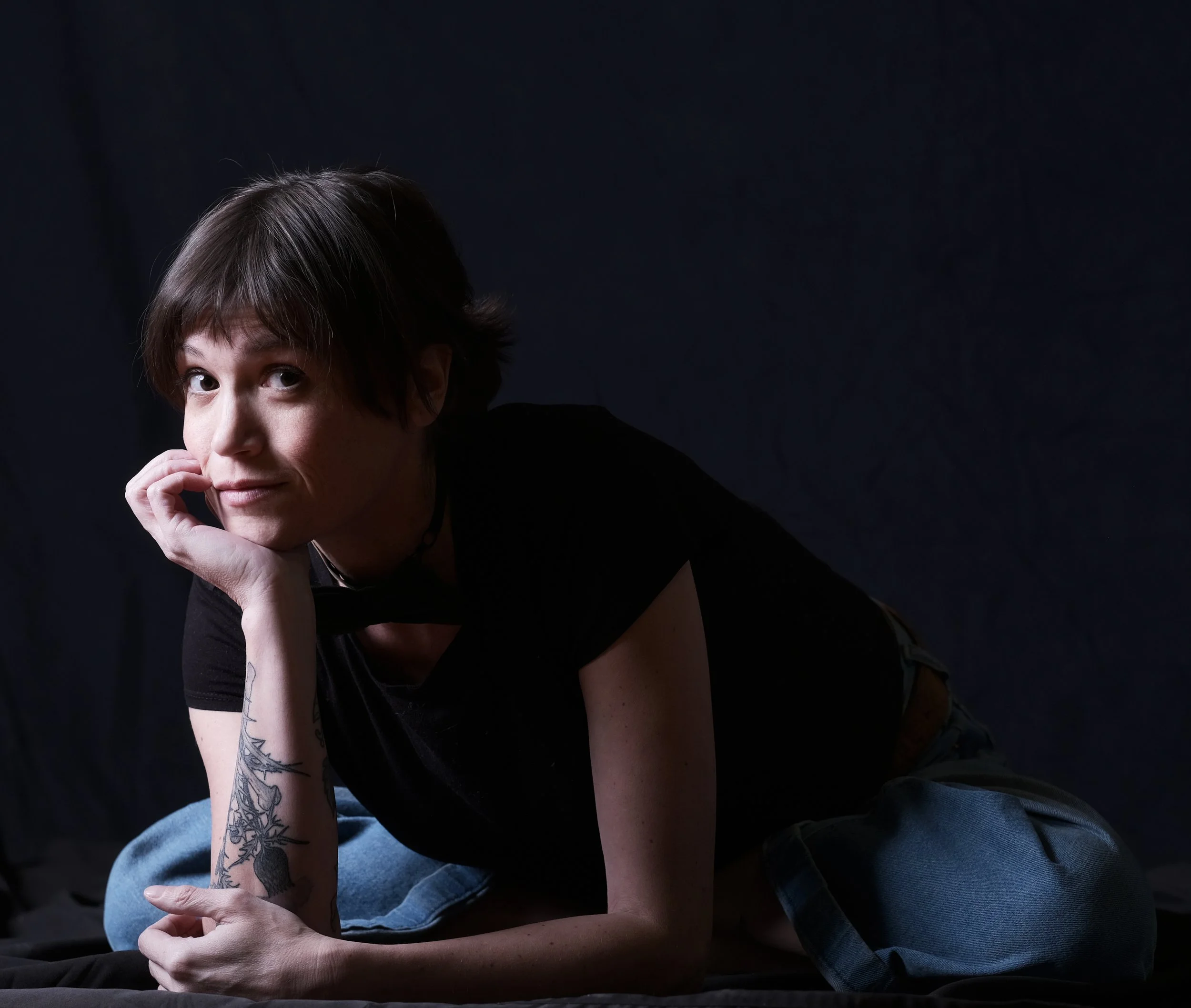 Person with short brown hair lying on their side, resting their head on their hand, wearing a black shirt and blue jeans, with a tattoo on their forearm, against a dark background.