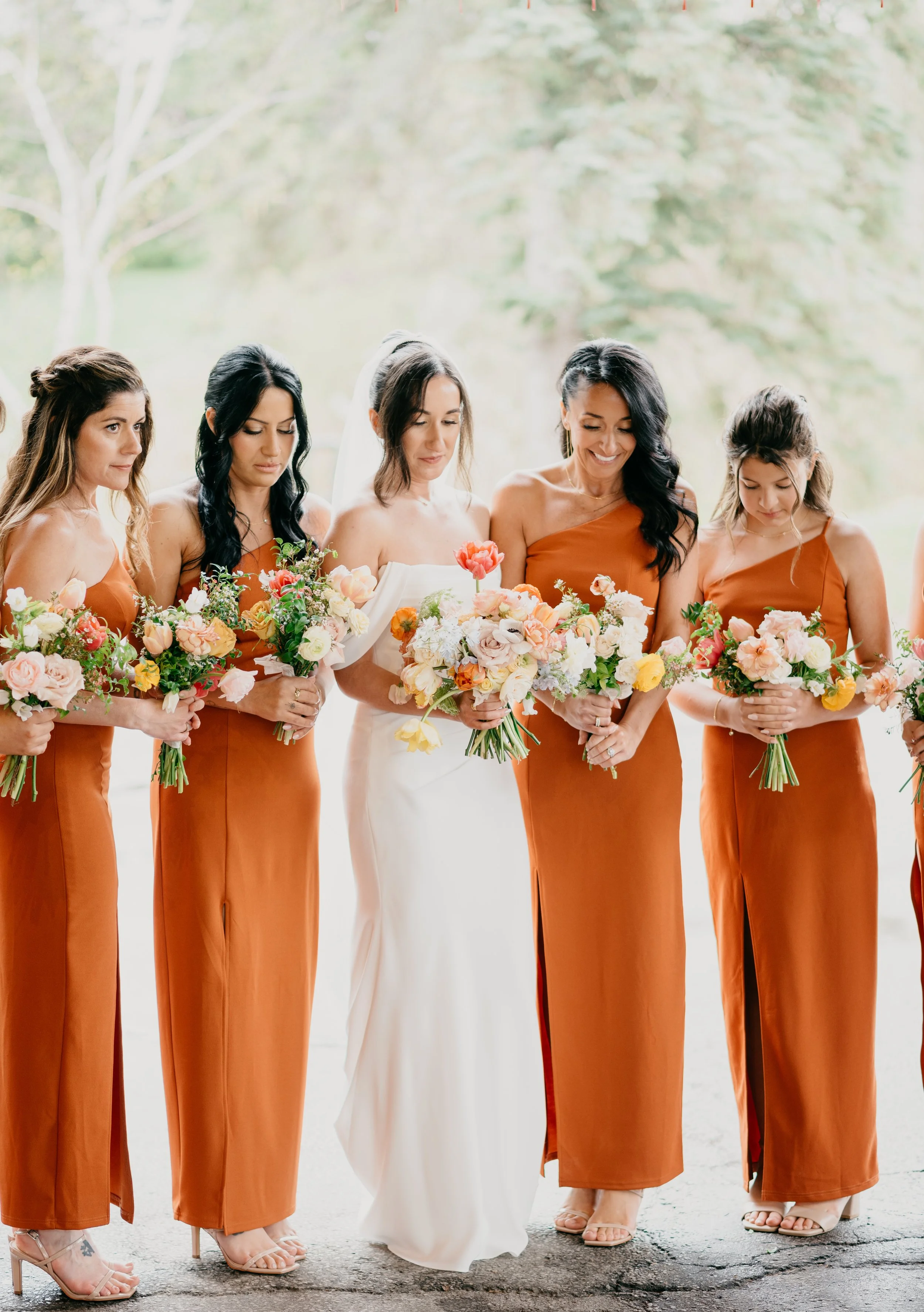 A bride in a white wedding dress holding a bouquet of flowers, standing among bridesmaids dressed in burnt-orange gowns, each holding a bouquet, outdoors with trees in the background.