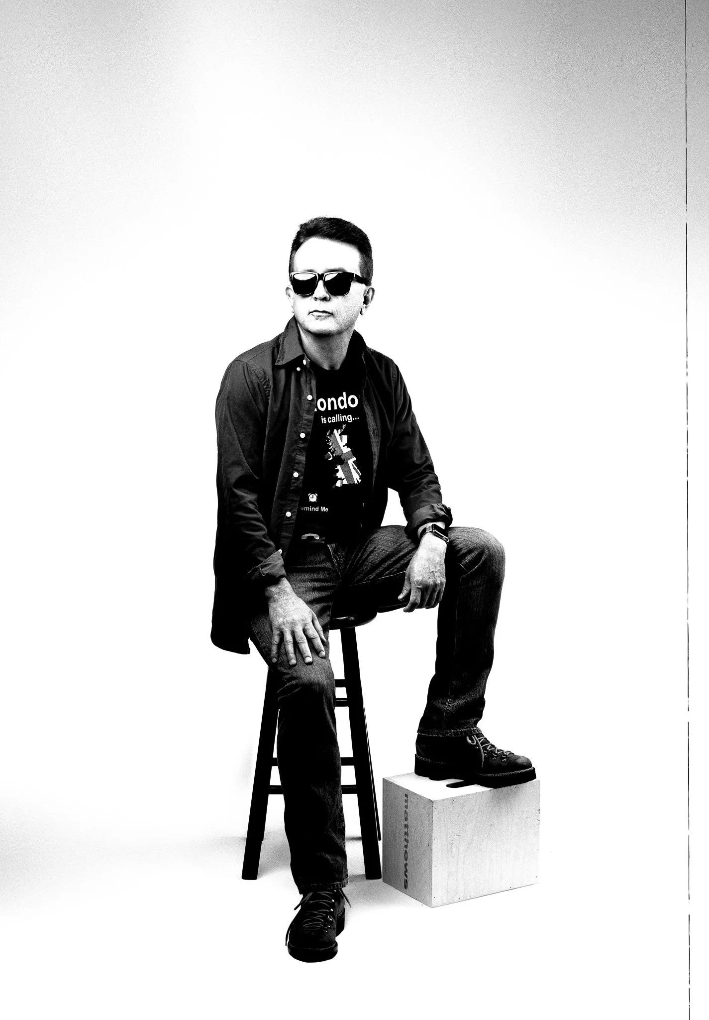 A man wearing sunglasses, a dark shirt, and jeans sitting on a stool with one foot on a wooden box, in a black-and-white studio portrait.