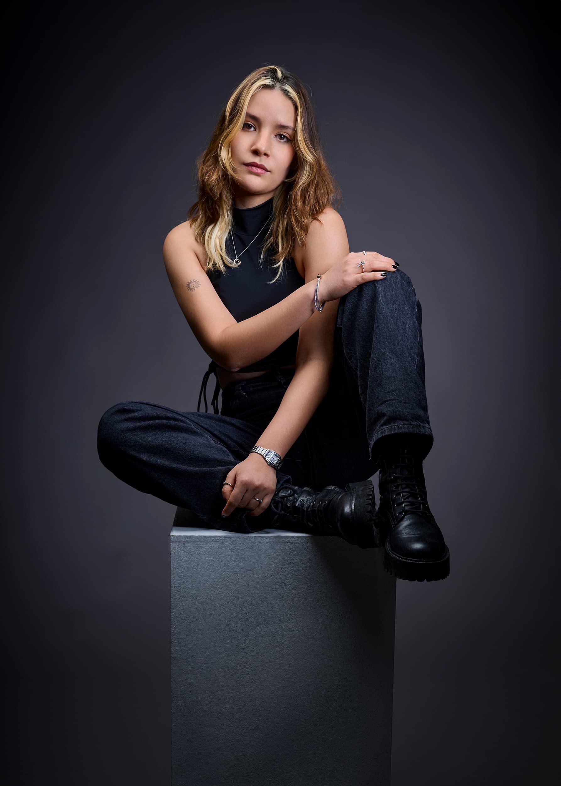 Creative portrait of a young woman with wavy blonde hair, sitting on a gray block against a dark gradient background, wearing a black sleeveless top and black pants, with black combat boots and silver jewelry, looking at the camera.