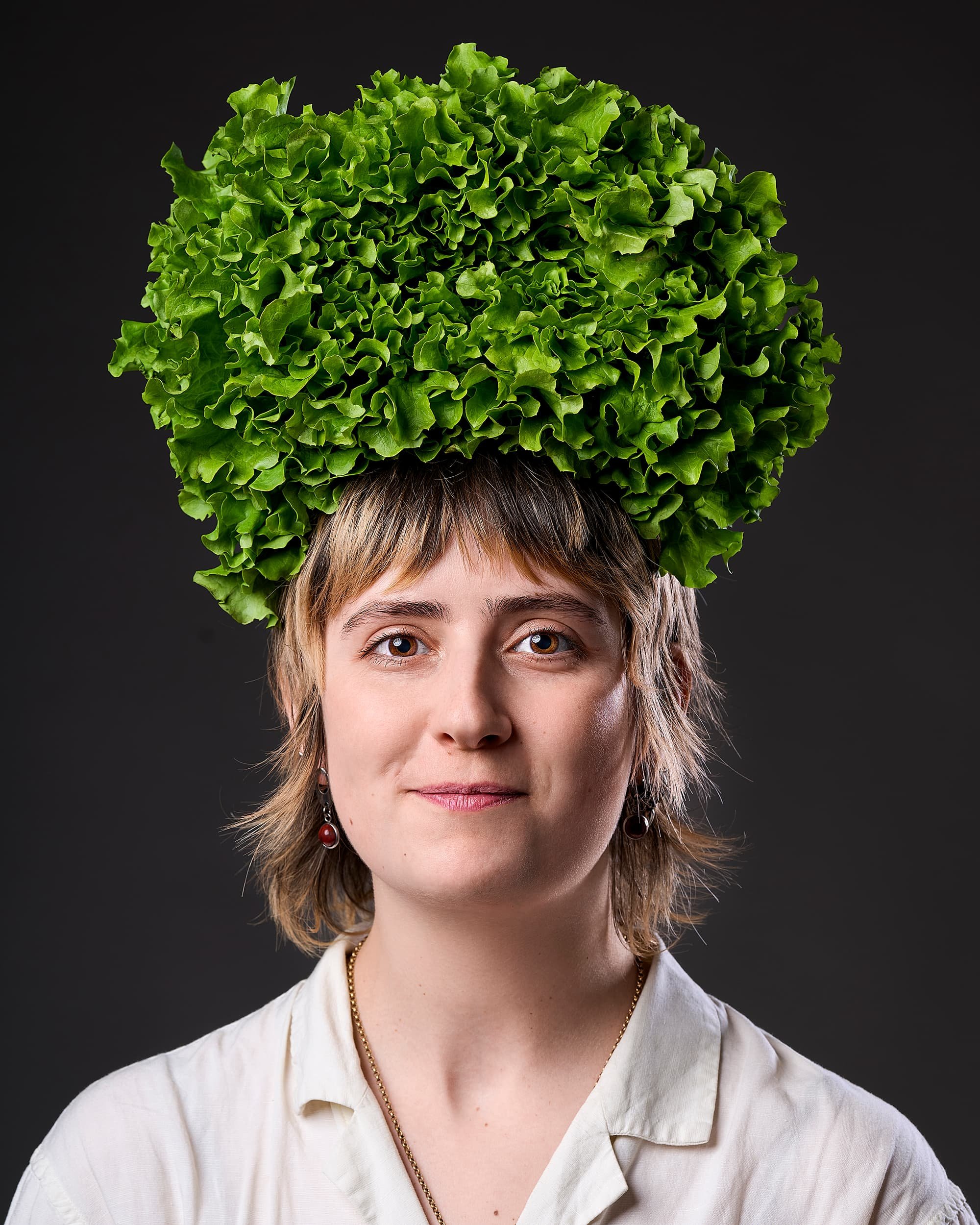 Creative portrait of a woman with short light brown hair wearing a white shirt and earrings, with a large head of green leafy lettuce on her head, against a dark background.