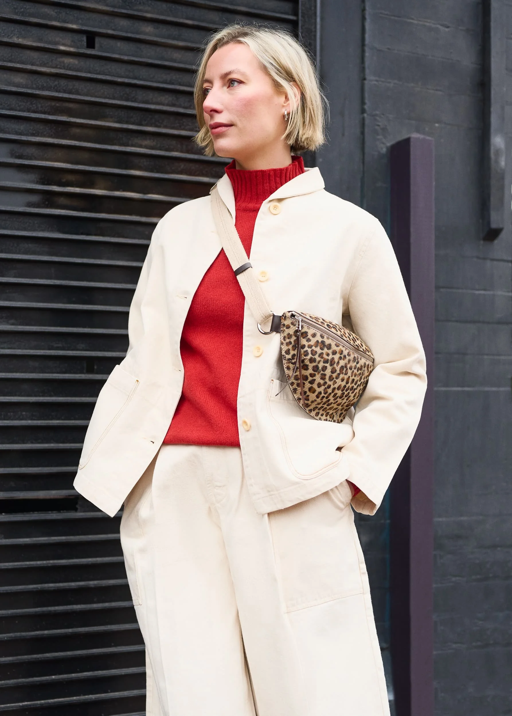 A woman with short blonde hair and earrings standing outdoors against a dark background, wearing a beige jacket, red sweater, and holding a leopard print bag.