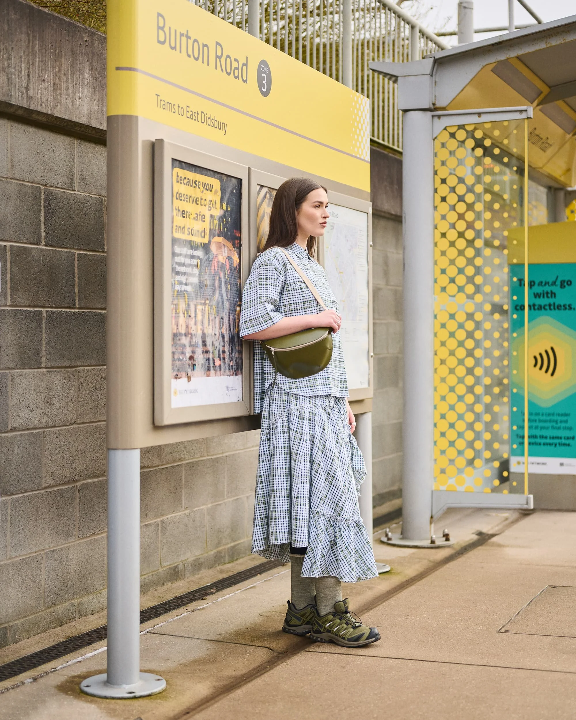 Fashion photograph shot on location of a young woman standing at a tram stop on Burton Road, wearing a checkered dress, gray socks, and hiking shoes, with a green purse over her shoulder, waiting at the tram stop displays advertisements.