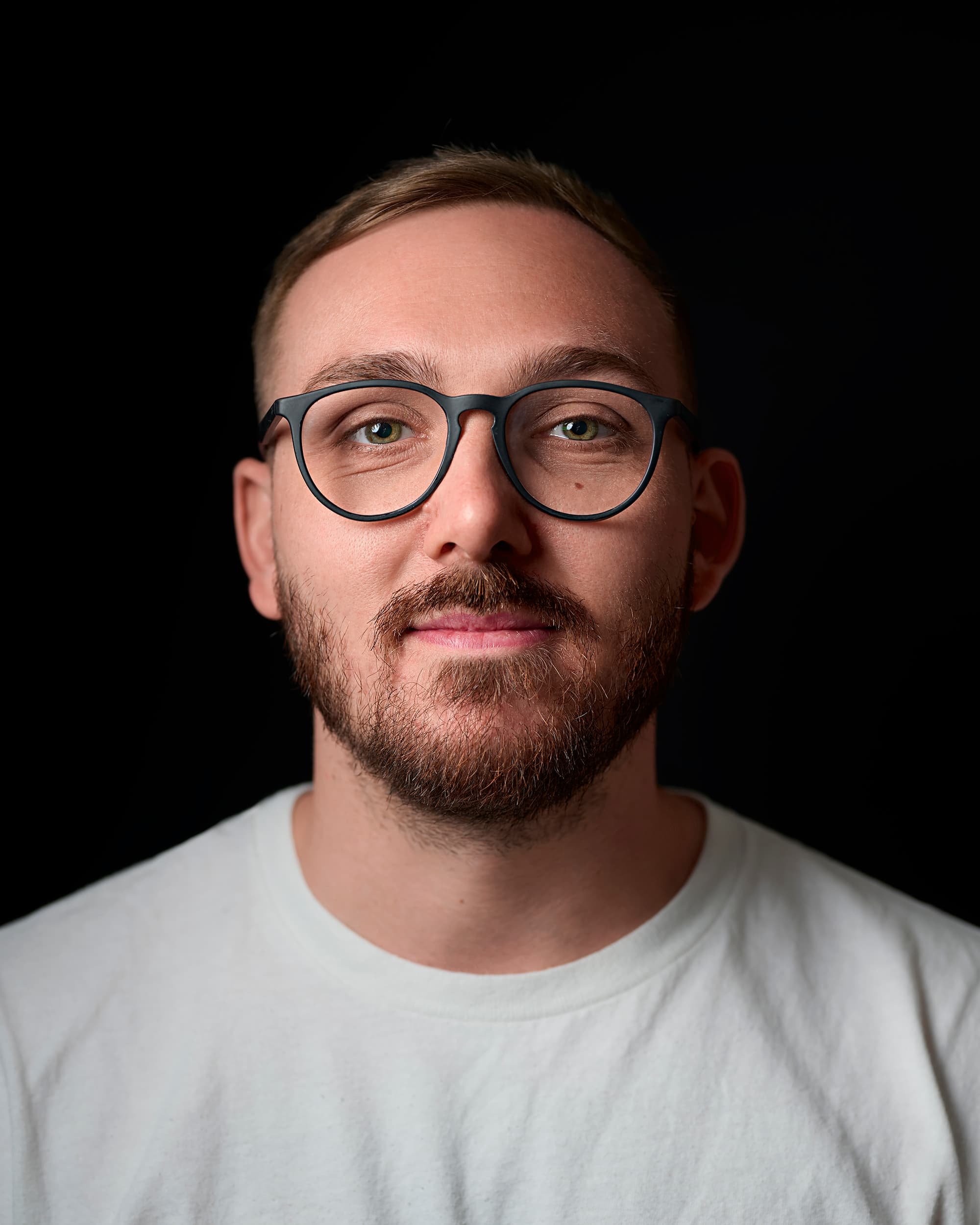 Close-up headshot portrait photograph of a young man with glasses, a beard, and light brown hair, wearing a white shirt against a black background.