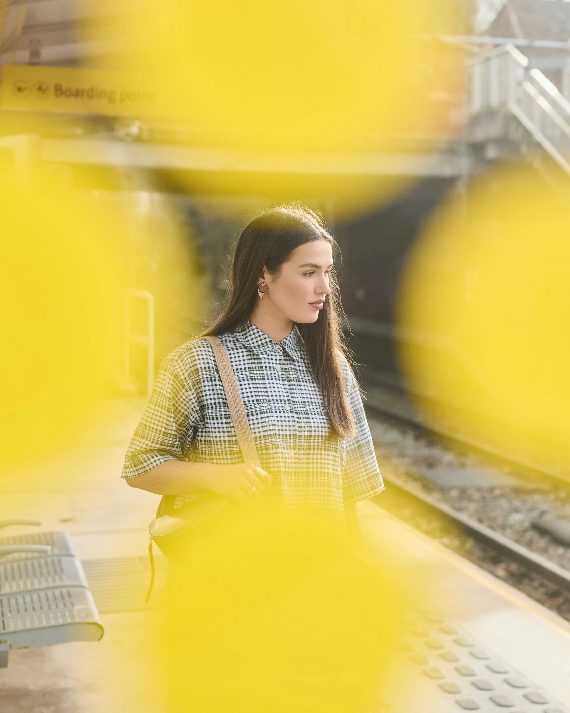 A young woman with long dark hair standing on a train platform, wearing a checkered shirt, carrying a beige shoulder bag, with yellow blurred out object in the foreground.