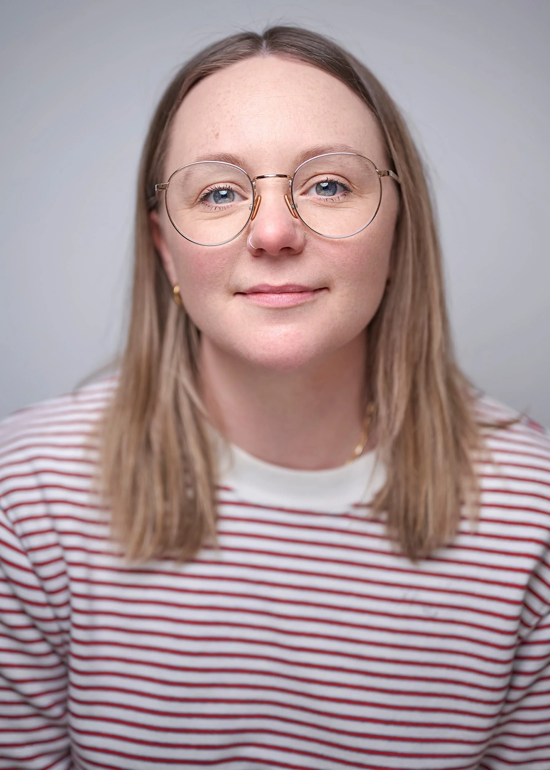 Professional portrait photograph of a woman with light brown hair, wearing glasses, a nose ring, and a white and red striped shirt, smiling softly against a plain light gray background.
