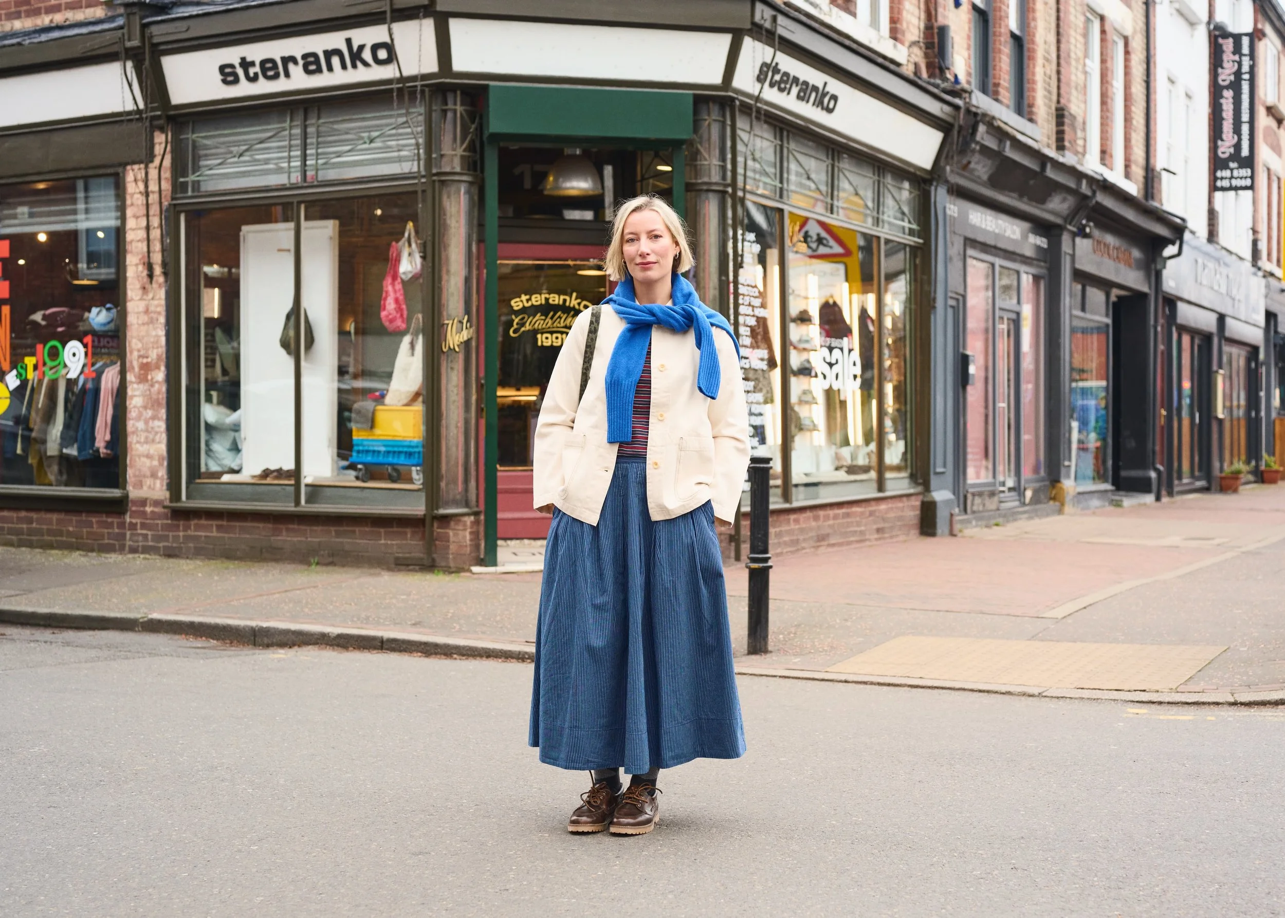 A young woman with blonde hair wearing a beige jacket, blue scarf, long blue skirt, and hiking boots standing on a city street in front of a storefront with large windows and a sign that says 'steranko'.