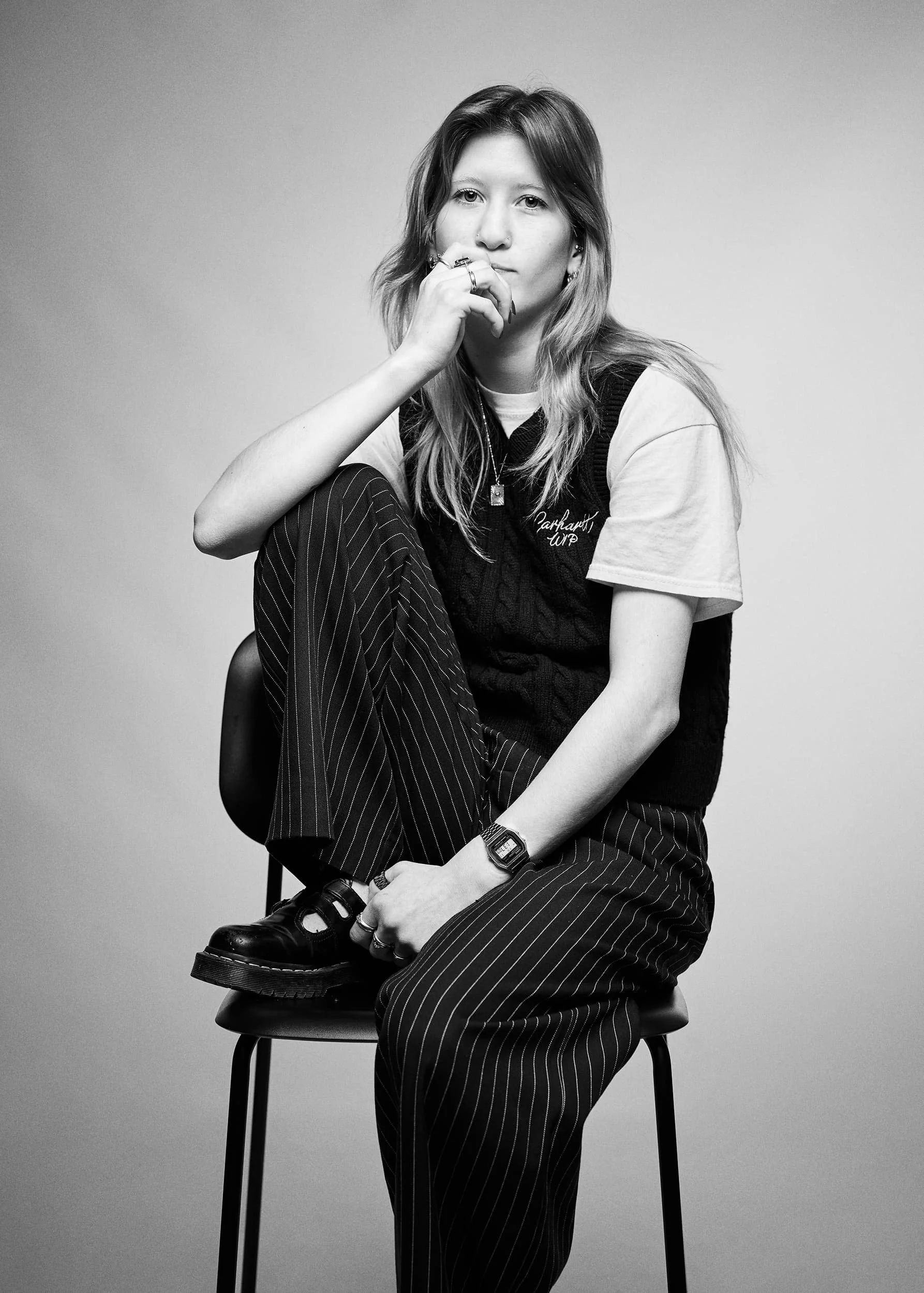 Portrait photograph if a young woman with long hair sits on a stool against a plain background. She is wearing a T-shirt, a sleeveless sweater vest, striped pants, and black shoes. She has one hand near her face and her leg resting on her knee.