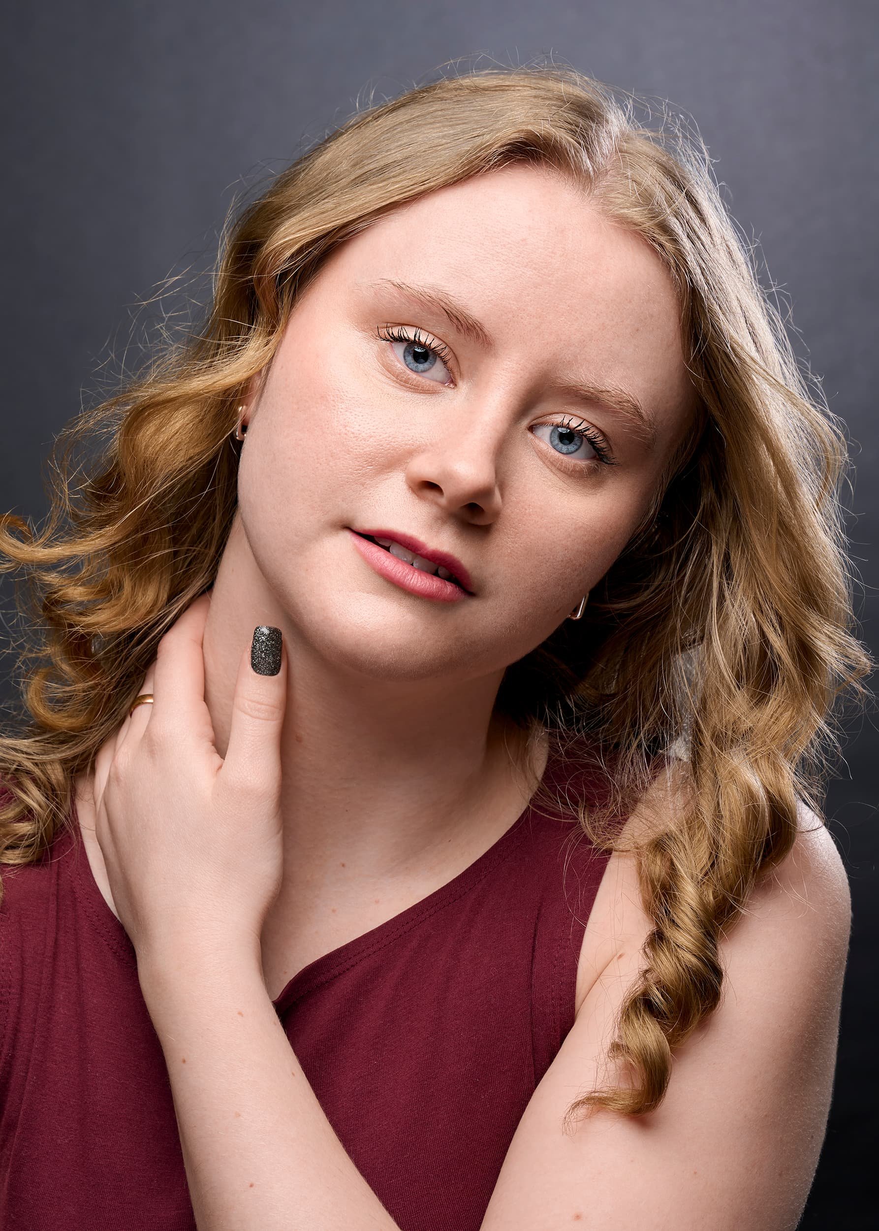 A young woman with wavy blonde hair, blue eyes, wearing a sleeveless maroon top, gently touching her neck with her right hand, against a dark grey background.