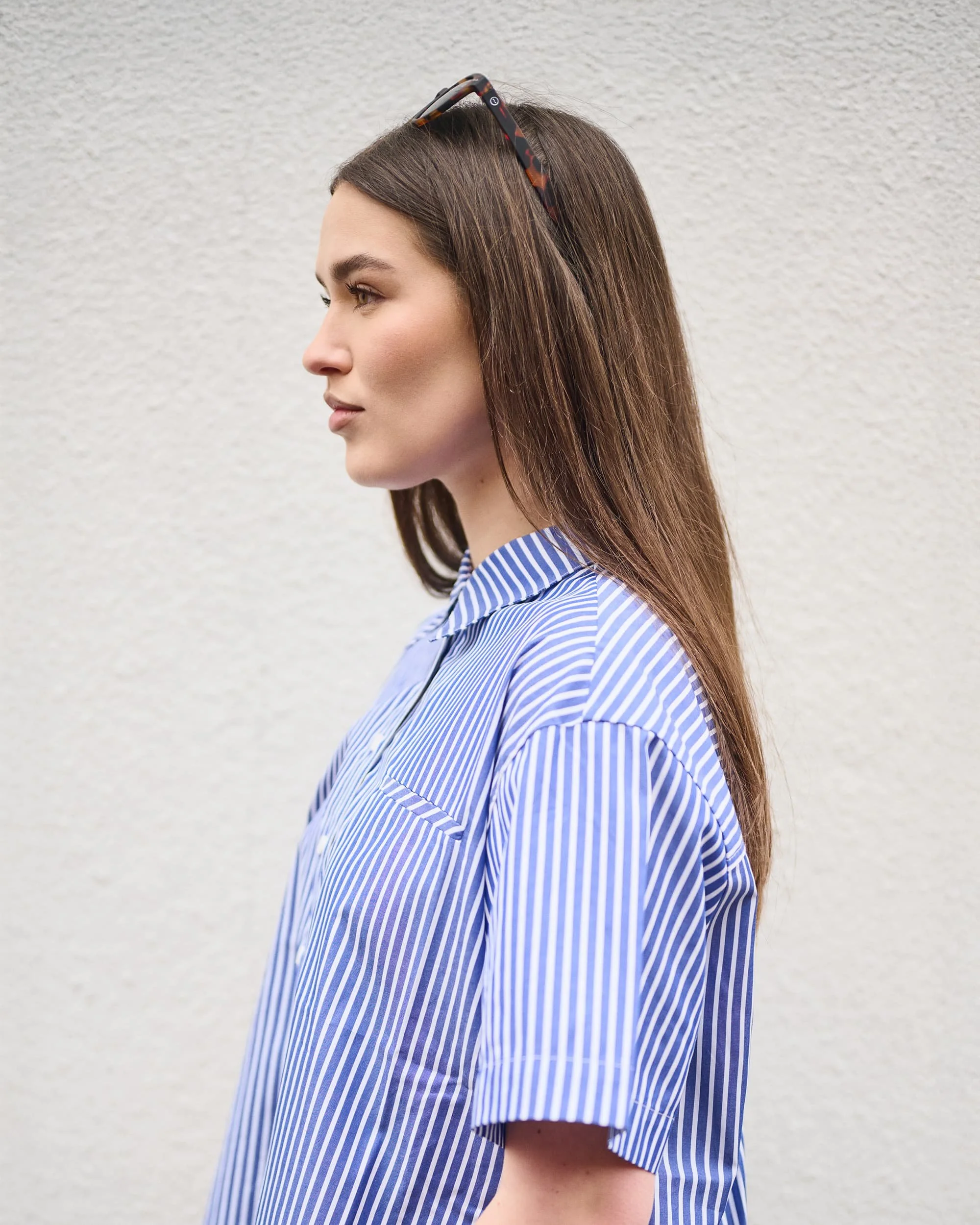 Profile of a woman with long brown hair, wearing sunglasses on her head and a blue and white striped shirt, standing against a plain white wall.