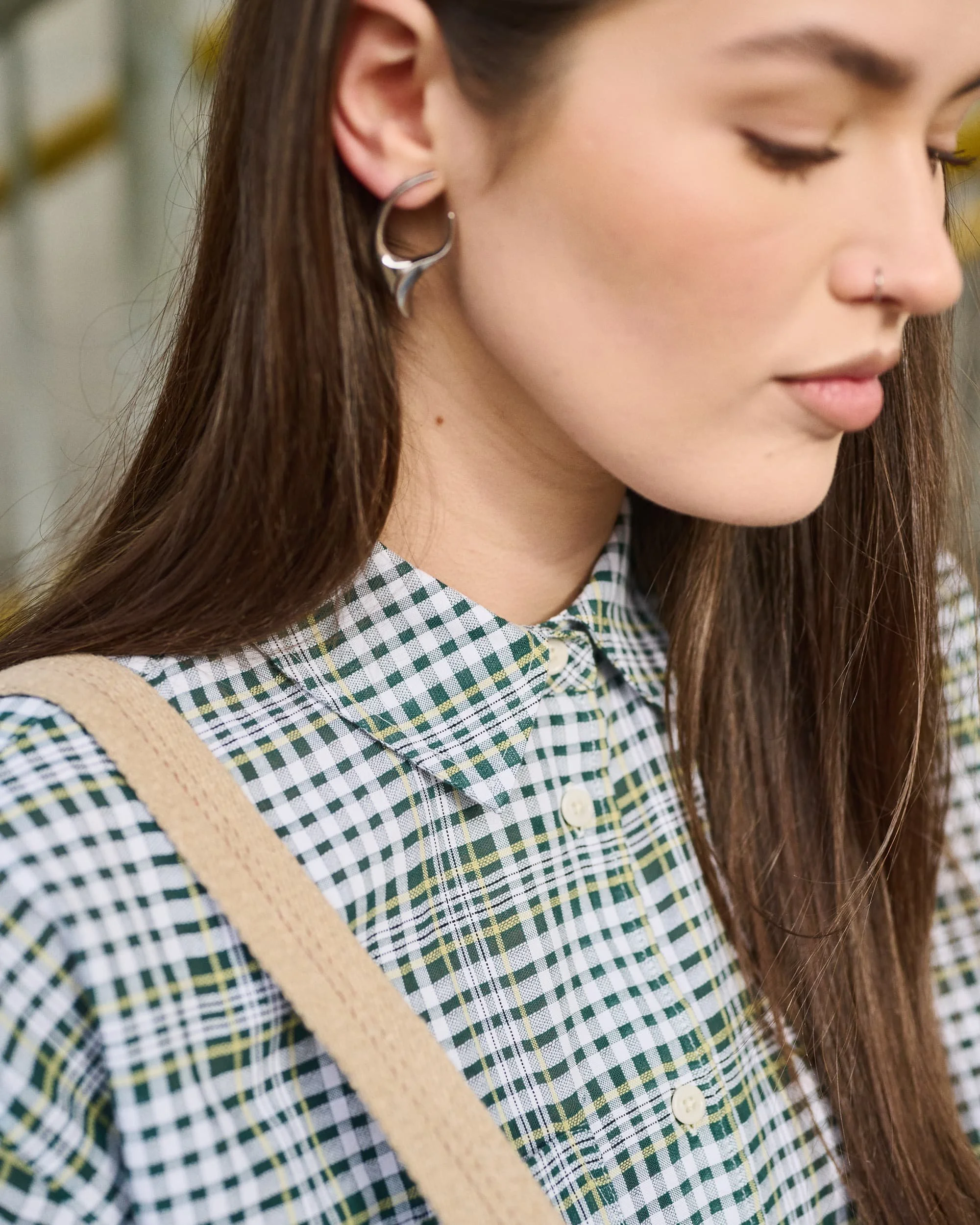 Close-up fashion photograph of a young woman with brown hair, wearing a plaid shirt, hoop earrings, and a nose piercing, looking down.