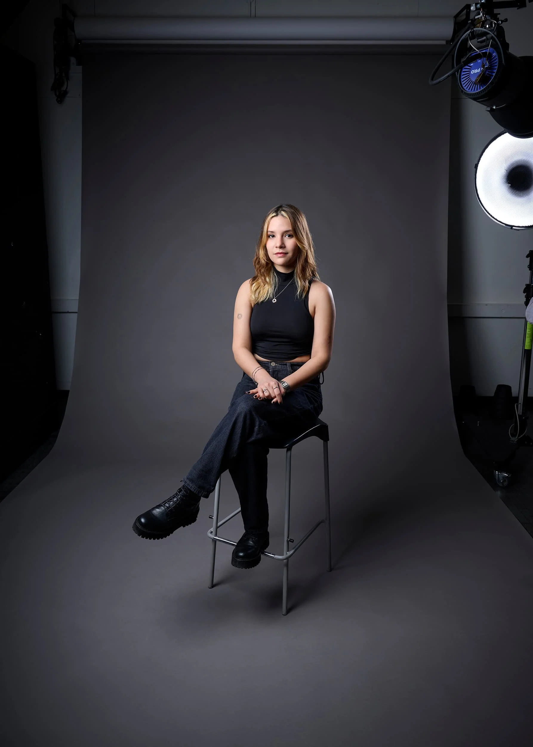 Portrait photograph of a young woman with light brown hair sits on a black stool in a photography studio with a dark gray backdrop, wearing a sleeveless black top and black pants, surrounded by professional studio lighting equipment.