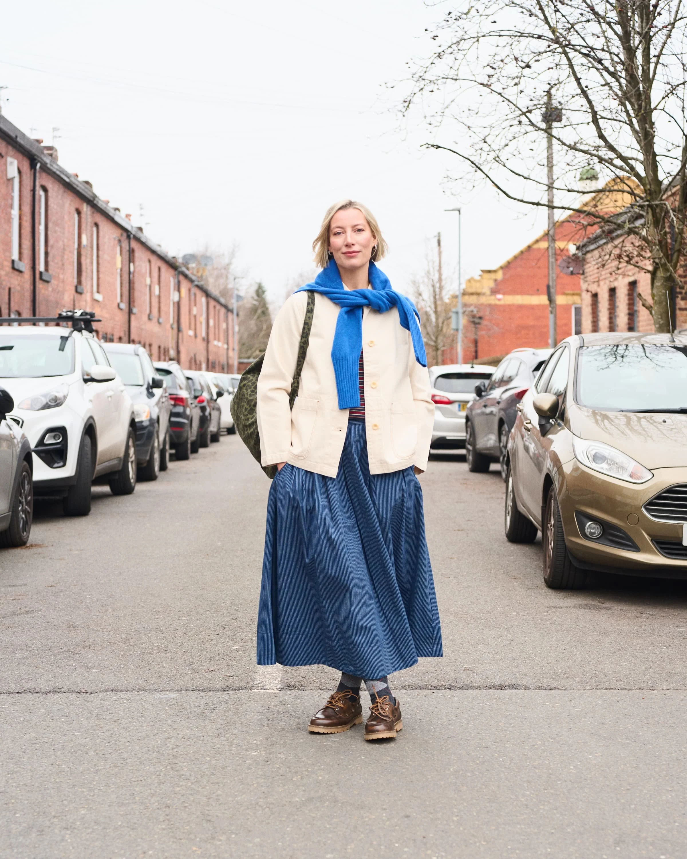 Editorial photograph of a woman standing in the middle of a city street with parked cars on either side, wearing a light-colored jacket, blue skirt, brown boots, and a blue sweater tied around her shoulders.