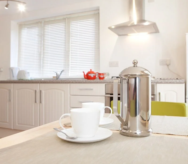 Kitchen with white cabinets, window with blinds, kettle, teapot, and cups on table, and stainless steel French press.
