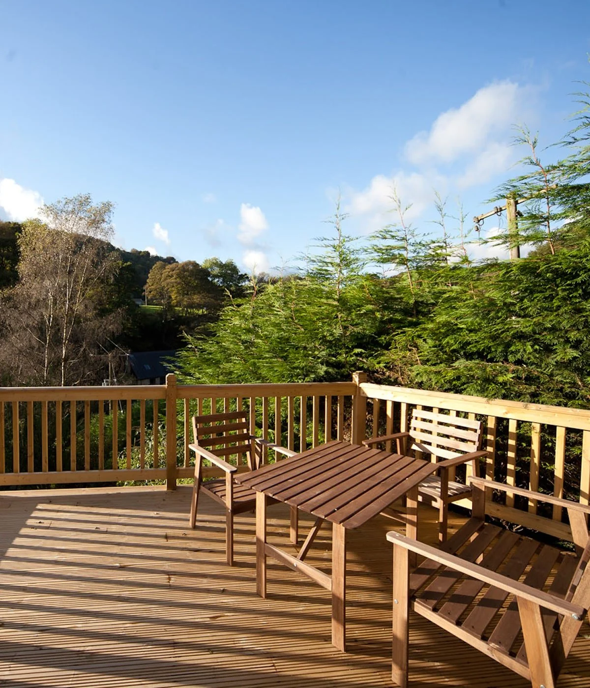 A wooden deck with outdoor chairs and a table, surrounded by trees under a clear blue sky.