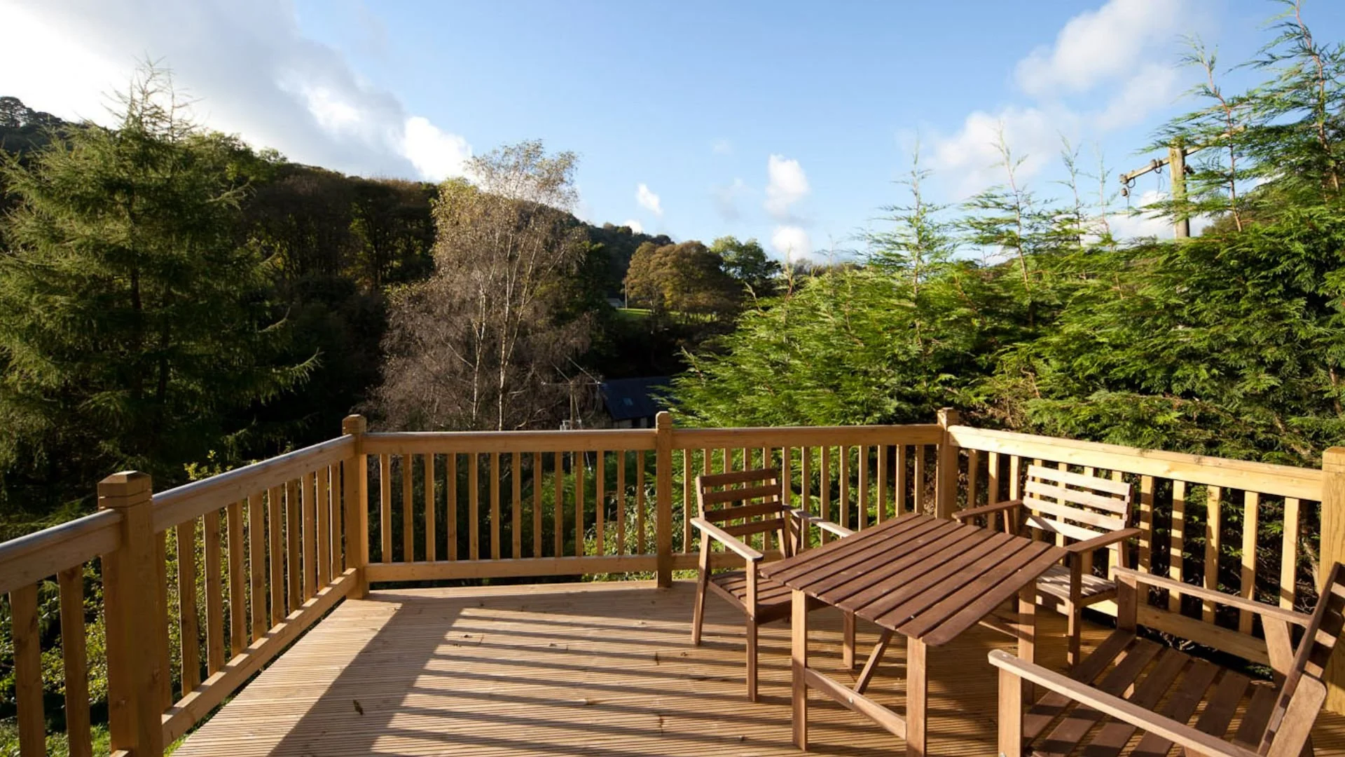 Wooden deck with four matching wooden chairs and a table, surrounded by green trees and a blue sky with clouds