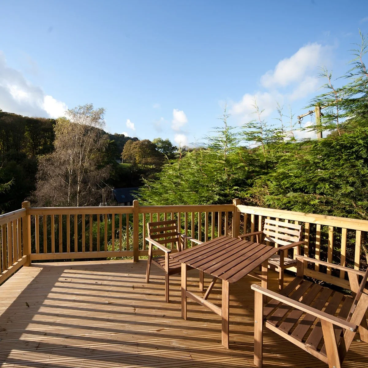 View of a wooden deck with outdoor chairs and table overlooking a green landscape with trees and a blue sky with clouds.