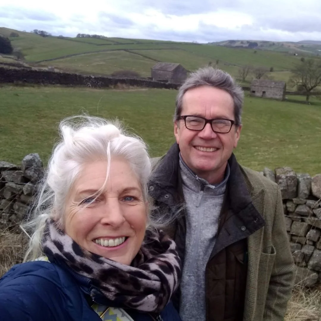 A smiling older woman with white hair and a scarf, and a smiling middle-aged man with glasses, outdoors in a rural green landscape with rolling hills, stone walls, and small farm buildings in the background.