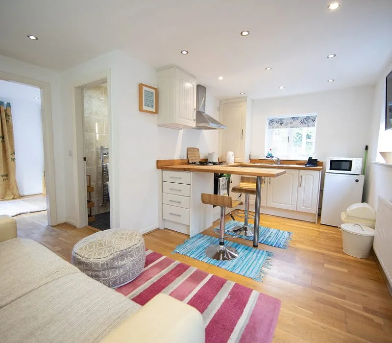 Open-concept kitchen and living room with wooden flooring, white cabinets, small bar stools at a breakfast bar, window with a patterned curtain, microwave, and various kitchen appliances.